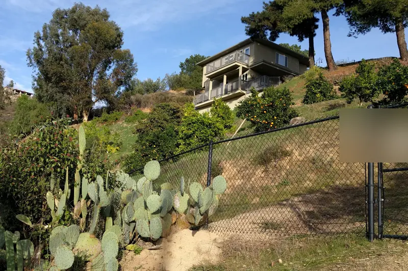 Hillside house above a terraced slope planted with cactus and fruit trees, seen from the road below