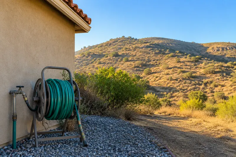 A long green garden hose on a reel beside an outdoor faucet on a stucco wall, a bare gravel strip at the base of the house and dry brown hills beyond