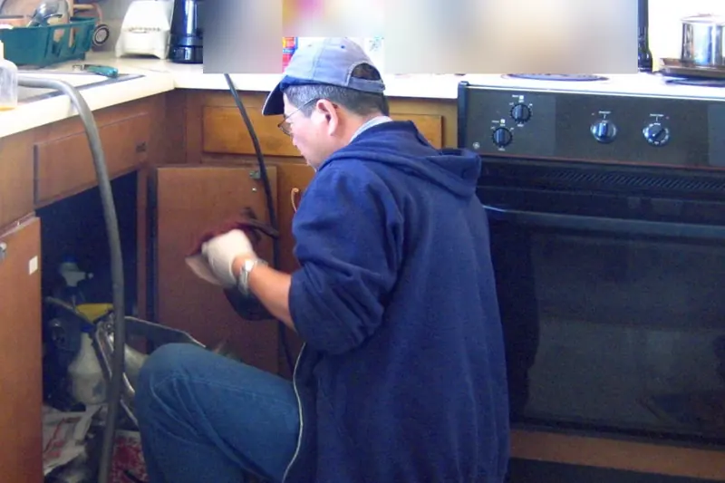 Plumber kneeling at an open kitchen sink cabinet, working on the drain and supply pipes underneath