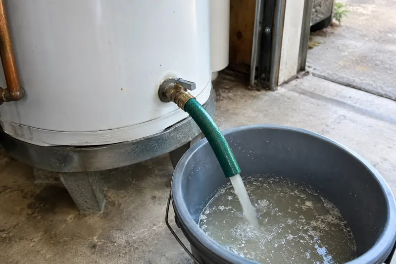 Garden hose screwed onto the drain valve at the base of a garage water heater, its other end draining into a bucket